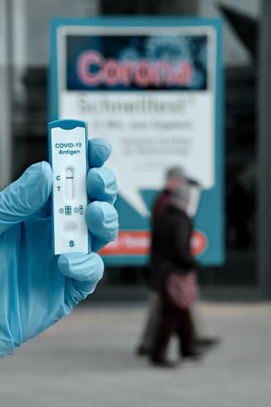 Hand in glove holding coronavirus test in front of banner for covid testing in Mitte. Schnelltest,...