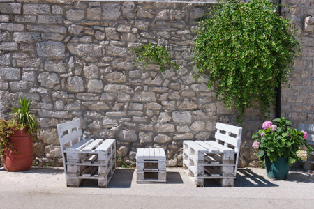 benches are situated beneath greenery in front of an historical stone wall between two flower pots
