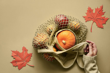 Autumn flat lay with female hands in olive green sweater and cotton string bag with orange pumpkins.