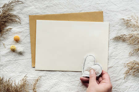 Textile flat lay with card, envelope, dry reeds, pampas grass. Hand holds soft heart. Off white cotton tablecloth background with cotton balls. Simple minimal flat lay with copy space, natural light.の写真素材