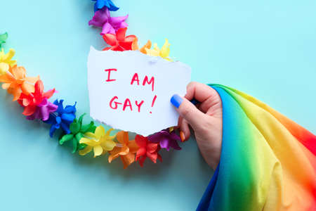 LGBTQ community pride. Text I am Gay on paper in hand with rainbow flag, flower garland and fake nails in colors of rainbow. Concept shot. Flat lay, top view on mint blue background.の写真素材