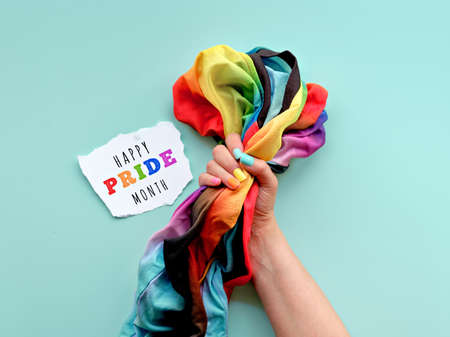 LGBTQ community pride. Hand with fake nails in colors of the rainbow holds rainbow t-shirt. Flat lay, top view, mint green background. Simple, minimal design in LGBT pride flag color elements.の写真素材