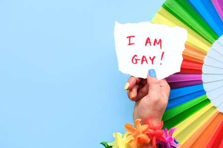 Text I am Gay on paper in hand with rainbow fan. LGBTQ community pride. Flower garland and fake nails in colors of rainbow. Flat lay, top view on mint blue background. Copy-space, text place.の写真素材