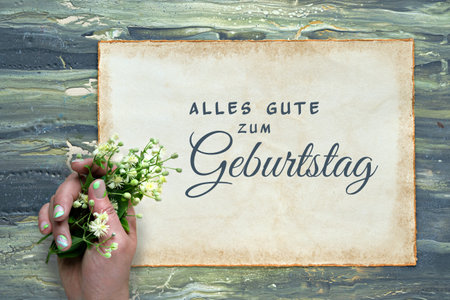Text Alles Gute zum Geburtstag means Happy Birhday in German. Greeting on aged patchment, beige paper page. Hand with pale yellow flowers. Flat lay on aged textured painted green brown background.の写真素材