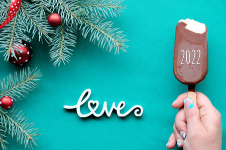 Happy New Year 2022. Hand with ice cream. WInter ackground on turquoise, green textile with wooden text Love. Top view on fir twigs decorated with red glass baubles.の写真素材