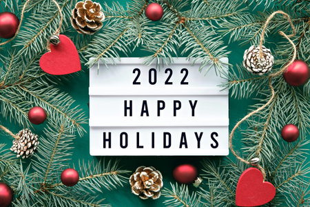 Light board with text 2022 Happy Holidays. Flat lay with fir twigs and Xmas decorations around light...