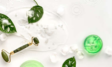 Green jade face roller on ice with ice cubes and exotic leaves. Monstera Adansonii Swiss Cheese leaves on off white background. Beauty facial massage with home made moisturizer concept top view.の写真素材