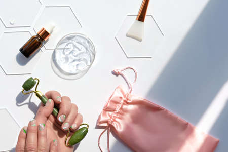 Hand with jade face roller and moisturizer gel in round glass dish. Home made beauty facial massage therapy. Off white flat lay with petri dish and glass hexagones. Natural light with long shadows..の写真素材
