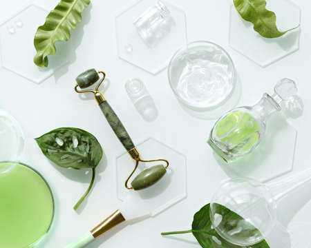 Moisturizer, green jade face roller on ice with ice cubes and exotic leaves. Monstera Adansonii leaves. Minimal flat lay on off white background. Facial massage with self made cosmetics.の写真素材