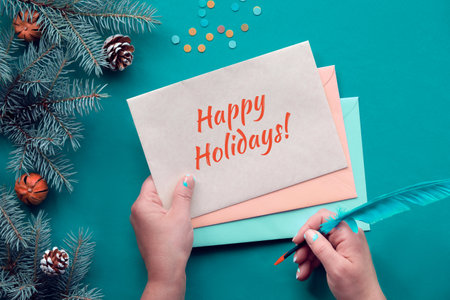 Traditional Christmas background with text Happy Holidays on postcards. Hands with greeting cards and quill. Top view on fir twigs decorated with red glass baubles, Xmas toys. Paper confetti.の写真素材