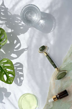 Moisturizer, green jade face roller with exotic leaves. Monstera Adansonii leaves. Sunshine, long shadows. Minimal flat lay on off white background. Facial massage background.の写真素材