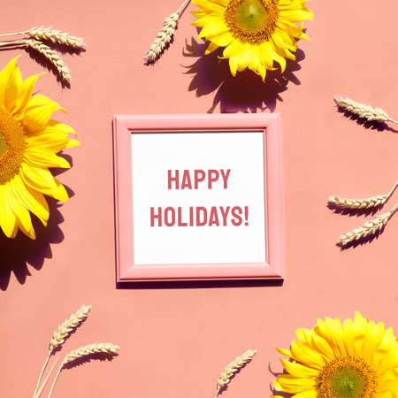 Text Happy Holidays in square frame. Flat lay with natural sunflowers, wheat ears. Sunlight, long shadows. Square composition with Autumn decorations on peach paper.の写真素材