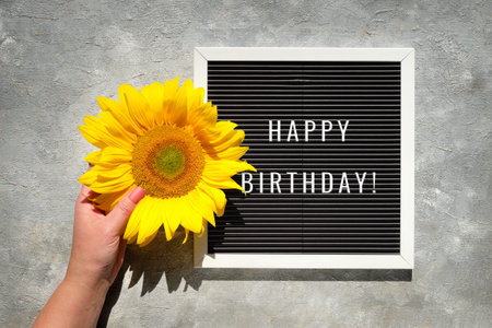 Text Happy Birthday on text board. Autumn natural decorations. Yellow sunflower in hand. Flat lay,...