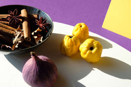 Card with Autumn colors. Purple fig, yellow quince fruits on vibrant fuchsia, magenta and white layered paper. Plate with pumpkin pie spices, cinnamon, anise. Direct sunlight, long shadows.の写真素材