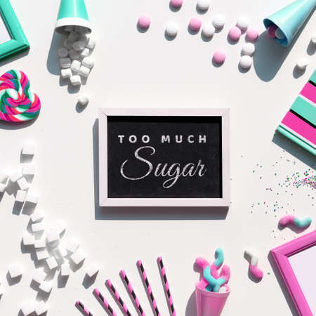 Text Too Much Sugar. Excess of sweets, chocolate, candy, sweet food. Sugar tax concept. Various tasty snacks in pastel colors, pink, mint green, on white background. Text on blackboard.の写真素材