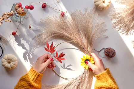 Hands making dried floral wreath from dry pampas grass and Autumn leaves. Hands in sweater with manicured nails tie decorations to metal ring. Flat lay on white background, sunlight with long shadows.の写真素材