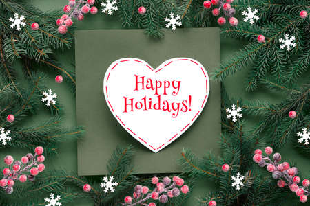 Green red Christmas background with text Happy Holidays on white paper heart. Natural Xmas twigs decorated with frosted berries and paper snowflakes. Flat lay, top view on green paper.の写真素材
