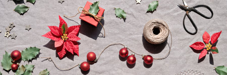 Christmas flat lay on natural beige linen textile. Hands decorate gift boxes. Red poinsettia, red glass baubles, winter decor. Dry rose tea leaves. Hemp cord, scissors, red and dark green poinsettia .の写真素材