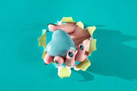 Giving Tuesday. Ceramic heart in wooden model hand through paper hole in torn cyan turquoise paper.の写真素材