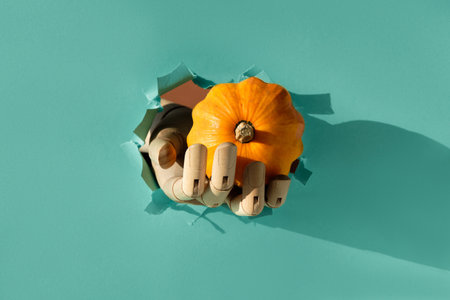 Pumpkin in wooden model hand through paper hole in torn mint green paper. Hello Autumn seasonの写真素材