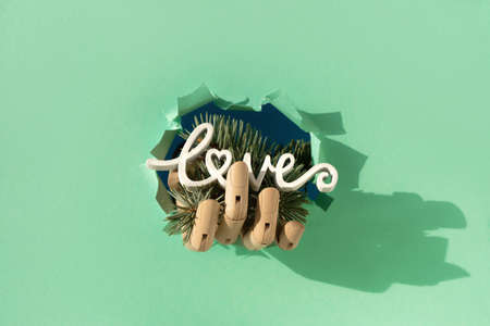 Merry Christmas, hand with fir twigs and wood word love through paper hole. Wooden model hand. Creative minimal concept background on mint green paper.の写真素材