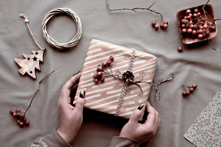 Eco friendly decorations - recycled craft wrapping paper, berry, wattle wreath, wood Christmas toys. Hands wrapping Xmas gift box with natural decor on beige textile tablecloth.の写真素材