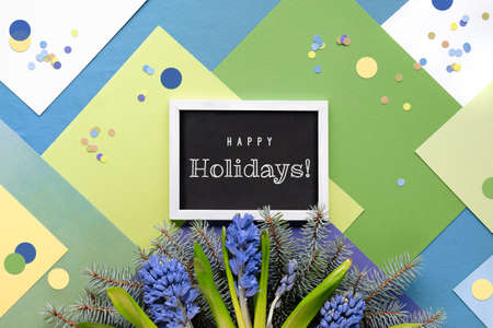 Text Happy Holidays on blackboard. Green layered geometric paper background with blue hyacinth flowers and fir twigs. Winter seasonal decorations. Paper circles, confetti.の写真素材