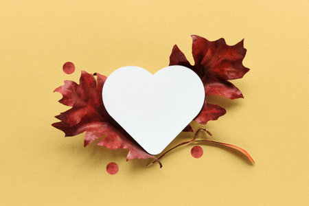 Blank paper heart shape with red autumn oak leave on yellow paper. Flat lay design element