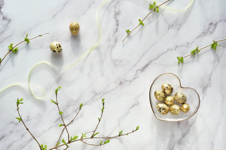 Golden quail eggs in glass heart shaped bowl. Twigs with young leaves on off white marble. Spring,...