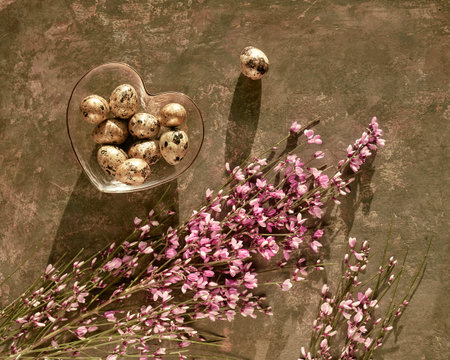 Spring Easter background with pink flowers, golden quail eggs in heart glass bowl on dark brown.
