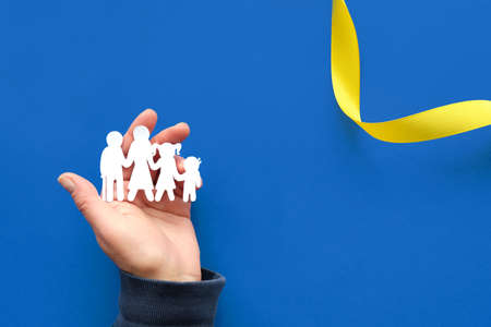 Helping hand holding Ukrainian family, paper silhouette. Flat lay, top view. Copy-space, text place on blue background with yellow ribbon.の写真素材