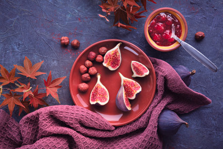 Autumntime background with fresh halved fig fruits on terracotta ceramic plate. Magenta towel and red maple Autumn leaves on dark purple board. Fig jam in ceramic bowl.の写真素材