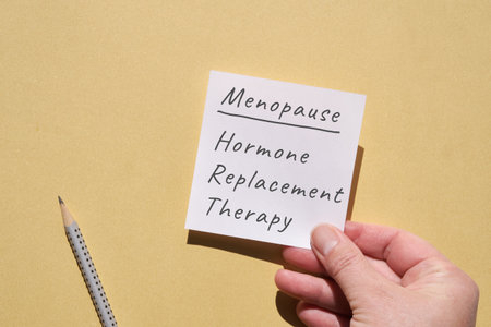 Hand hold paper page with text Menopause, hormone replacement therapy. Top view on yellow monochromatic paper background with pencil. Writing on blank square post it note.の写真素材