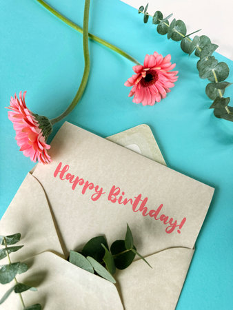 A pink envelope with a green birthday card placed inside, with pink gerbera flowers on white tableの写真素材