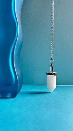 White crystal pendant, pendulum on a chain, healing amulet, spiritual energy detector on wavy blue paper backgroundの写真素材