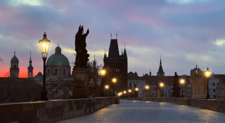 Dawn in Prague: Charles Bridge with pink-purple clouds, statue silhouettes, and glowing street lightsの写真素材
