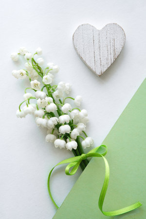 White lily of the valley flowers tied with a green ribbon placed on a white surface with a wooden heart and green paper, creating a delicate composition.の写真素材