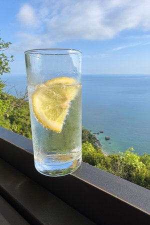 Glass of sparkling water with a lemon slice placed on a railing, offering a scenic view of the Adriatic Sea and lush greenery along the coastal road in Montenegro.の写真素材