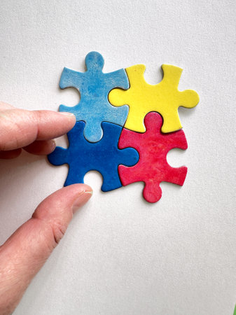 Close-up of a hand arranging blue, yellow, red, and green puzzle pieces on a white surface. Represents Autism Awareness Day in April and inclusivity.の写真素材