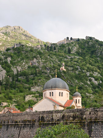 Orthodox church with distinctive domes surrounded by historic stone walls in Kotor, Montenegro. Green hills and ancient fortress ruins in the background.の写真素材