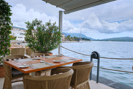 Scenic outdoor dining setup featuring wicker chairs and a wooden table with tableware, overlooking the tranquil waterfront in Porto Montenegro, Montenegro.の写真素材