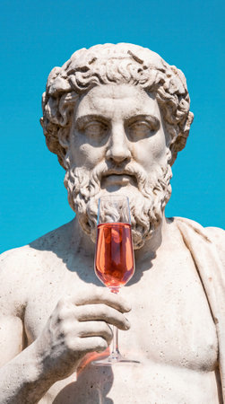 Classical marble statue holds a modern glass of rose wine against a clear blue sky, juxtaposing ancient sculpture with contemporary leisure and drink culture. Ample empty sky provides space for text and design elementsの素材