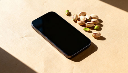 A sleek black smartphone lies on a beige surface alongside a small pile of pistachio nuts. The scene is illuminated by warm natural sunlight, creating soft shadows.の素材