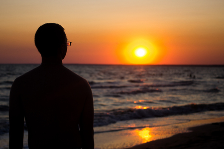 Silhouette of a young man with glasses looking at the sunset in the oceanの写真素材