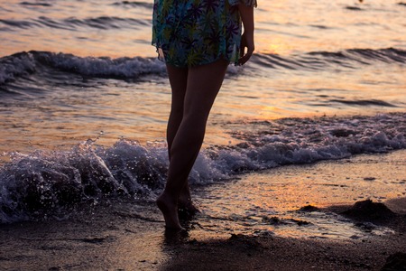Closeup of a bare feet of a girl walking on the beach at sunset, with a wave edge, foaming gently underneath them, toned colorsの写真素材