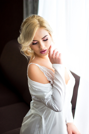 Beautiful blonde girl sitting on a sofa dressed in a white negligee holding a wedding bouquet, sending an air kiss in the background many colors.の写真素材