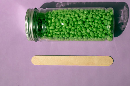 Beautiful green wax granules for depilation in a transparent jar, and wooden spatulas on a light...