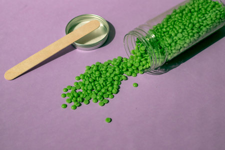 Beautiful green wax granules for depilation are poured from a transparent jar on a light purple...