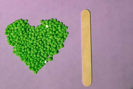 Beautiful green wax granules with Aloe extract for depilation are poured on a light purple background, in the shape of a heart. wooden spatula. Epilation, depilation, removal of unwanted hairの写真素材