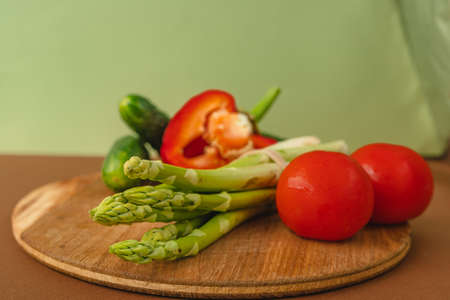 Vegetables lie on a wooden board: tomatoes, asparagus, cucumbers, red bell peppers. brown, light green background. place for text.の写真素材